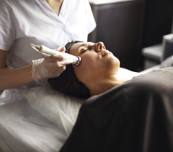 Cosmetician using modern machine for radiofrequency therapy on face of female client